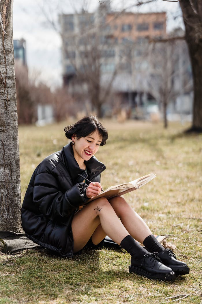 The Art of Drawing Readers In: Your attractive post title goes here Full body of smiling Asian female student taking notes in workbook while sitting on grassy lawn against residential buildings in town